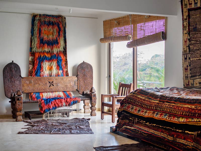 A vibrant room with tribal rugs & carpets, carved wooden furniture, and a bed draped in patterned fabrics beside large windows - Beautiful Homes
