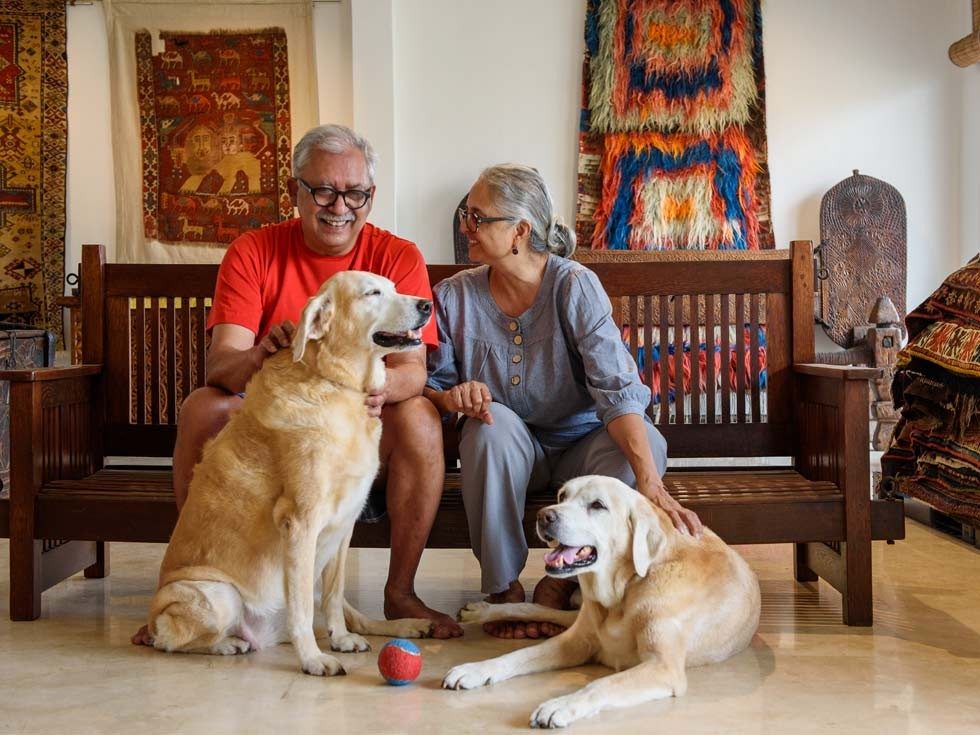 An elderly couple sits on a wooden bench with two dogs, surrounded by various patterned rugs - Beautiful Homes