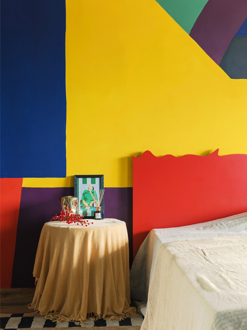 A vibrant room with a bed and a round table, set against a backdrop of boldly painted geometric walls – Beautiful Homes
