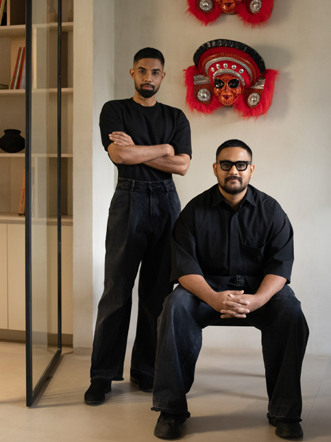 Arun Shekar and Mohammed Afnan of Humming Tree studio, known for their playful and bold designs, pose against a wall adorned with traditional masks – Beautiful Homes