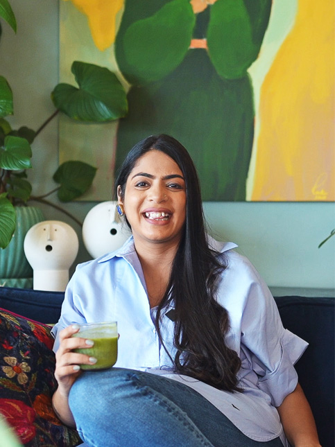 Devika smiles in her vibrant living room, surrounded by potted plants, eclectic curios, colourful textiles, and framed art – Beautiful Homes