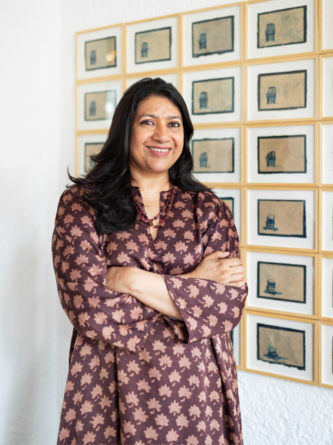 Pavitra Rajaram, in a patterned tunic, smiles confidently before a grid of framed artworks – Beautiful Homes