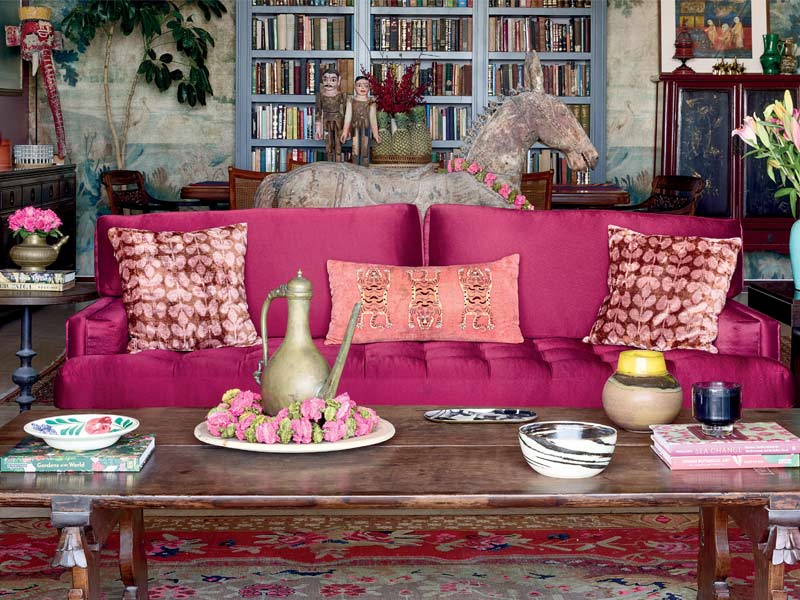 Pavitra Rajaram's living room features a raspberry velvet sofa, layered with vibrant cushions, a patterned rug, and a towering bookshelf – Beautiful Homes