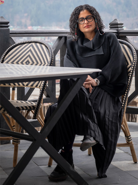 Shabnam Gupta, clad in black with colourful braided hair, sits thoughtfully at an outdoor table – Beautiful Homes