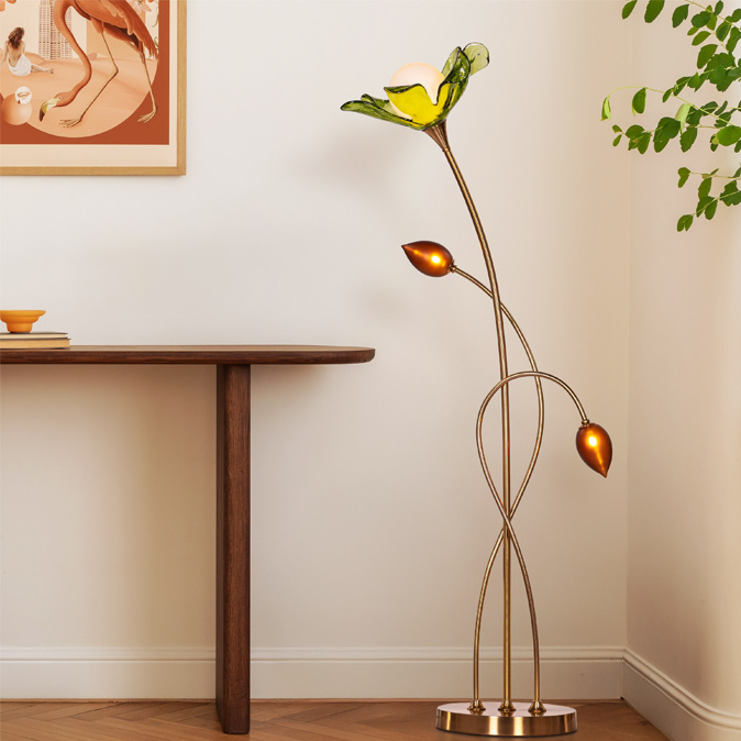 A whimsical, flower-inspired floor lamp with a yellow bloom and two amber buds illuminates a minimalist room with a wooden table and framed art – Beautiful Homes