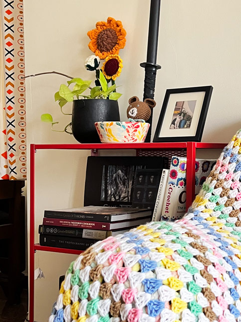 Colourful granny square crochet blanket draped on a modern grey sofa with a red shelf, books, and crochet flowers – Beautiful Homes