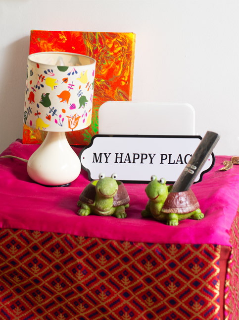 Cheerful tabletop décor with “My Happy Place” sign, floral lamp, vibrant art, and turtle figurines on a bright cloth – Beautiful Homes
