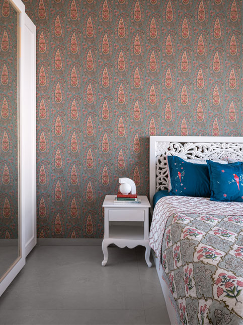 A guest bedroom with a striking gray wallpaper featuring a red and blue paisley pattern, a bed with a simple white bedside table - Beautiful Homes
