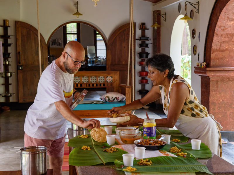 A man and a woman are standing at a wooden table covered with banana leaves, serving food for a meal - Beautiful Homes