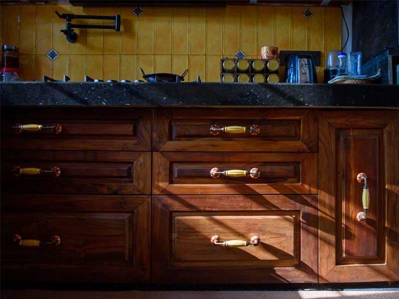 Rustic dark wood kitchen cabinets with decorative handles, black granite countertop, and a yellow tiled backsplash highlighted by sunlight - Beautiful Homes