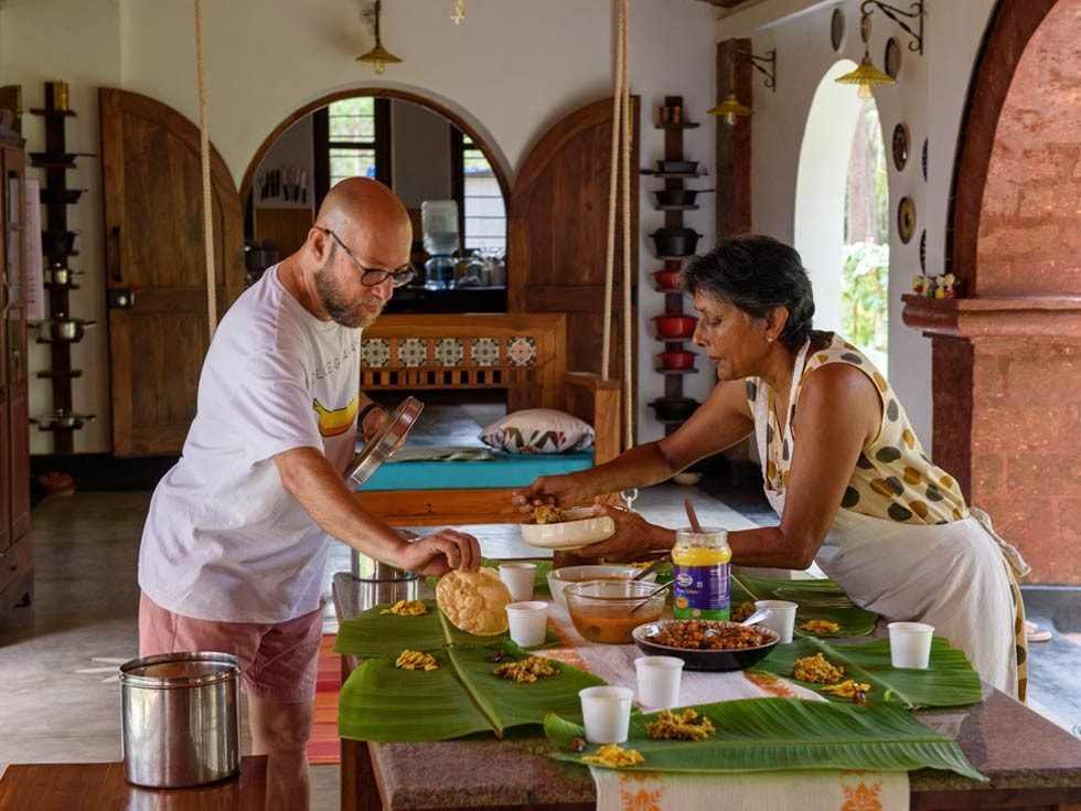 A man and a woman are standing at a wooden table covered with banana leaves, serving food for a meal - Beautiful Homes