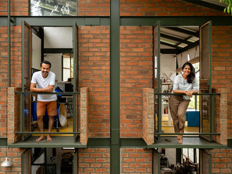 Designers Anindya and Garima Roy stand on small balconies of a red brick and steel framed building, looking out and smiling at the viewer – Beautiful Homes