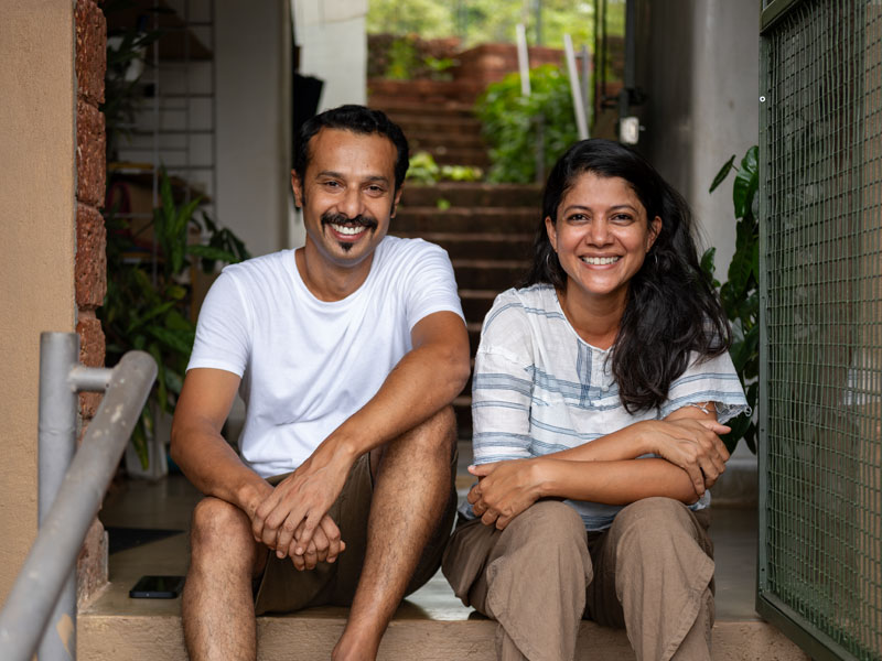 Designers Anindya and Garima Roy stand on small balconies of a red brick and steel framed building, looking out and smiling at the viewer – Beautiful Homes