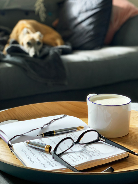 Close-up of a notebook, pens, and glasses on a tray with a dog relaxing on a couch – Beautiful Homes