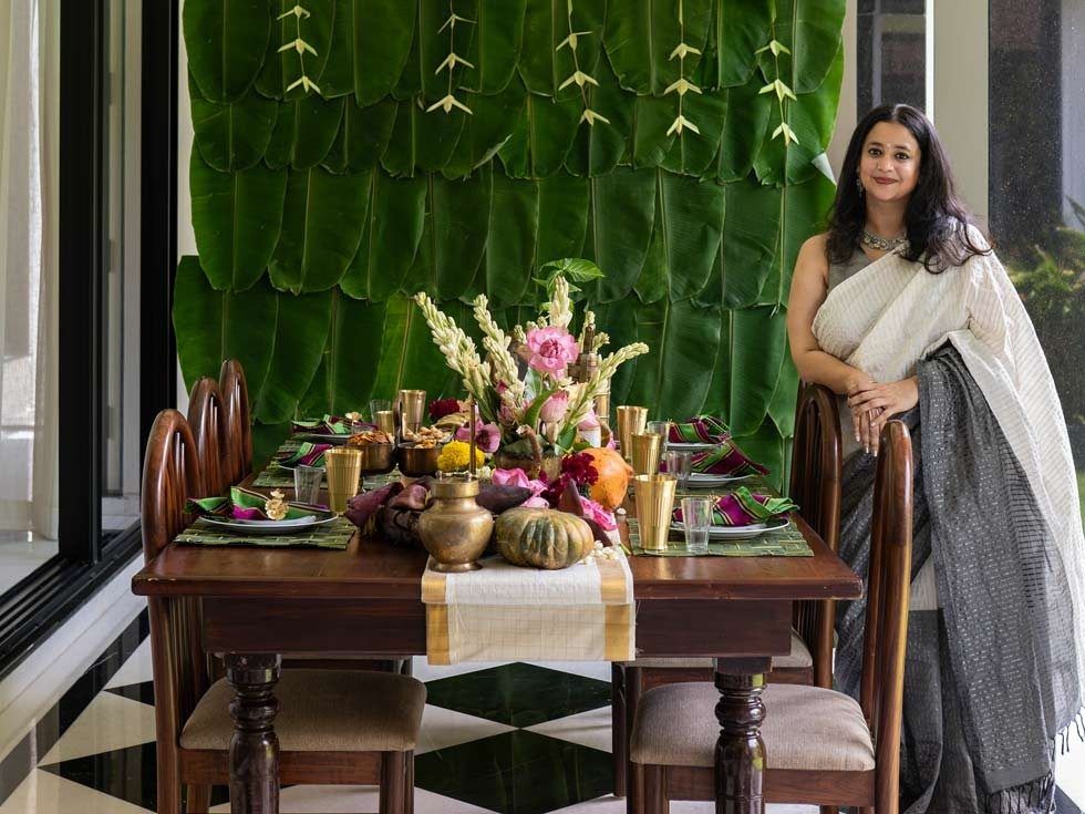 Varsha Menon standing beside a festive Onam tablescape at her Chennai home, featuring banana leaves, brass accents, flowers and seasonal produce - Beautiful Homes