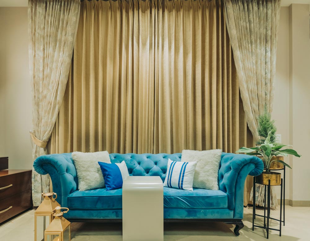 Livign room with two curtain designs, a blue sofa, golden elements and blue cushions