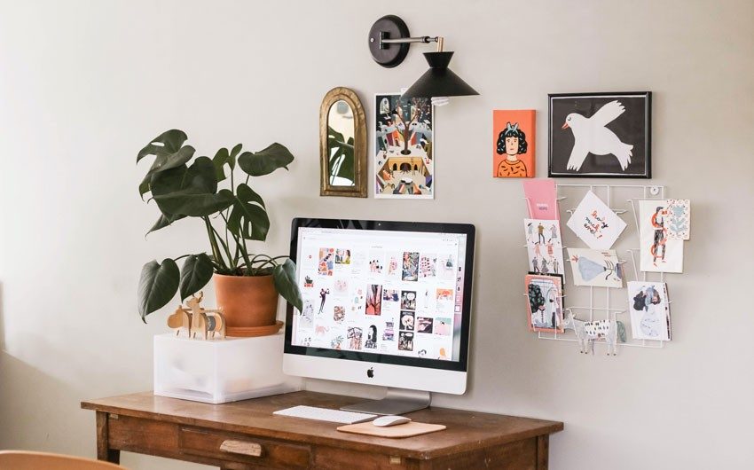 A wooden desk with a computer on it, a planter, a desk organiser and on the wall behind it are frames of artwork hung on it