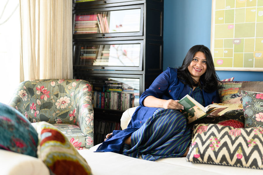 Designer Pavitra Rajaram sitting on a bed with colourful cushions, with a floral chair, black bookshelf and blue walls - Beautiful Homes