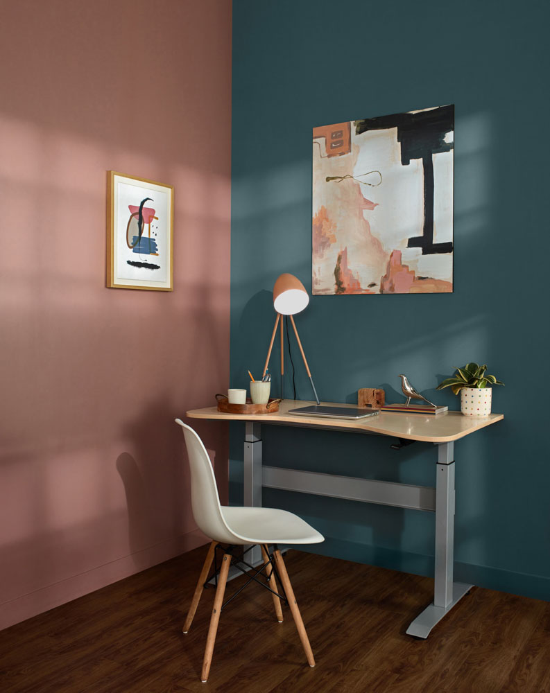A study table placed in a corner of a room with a table lamp and artworks on the walls