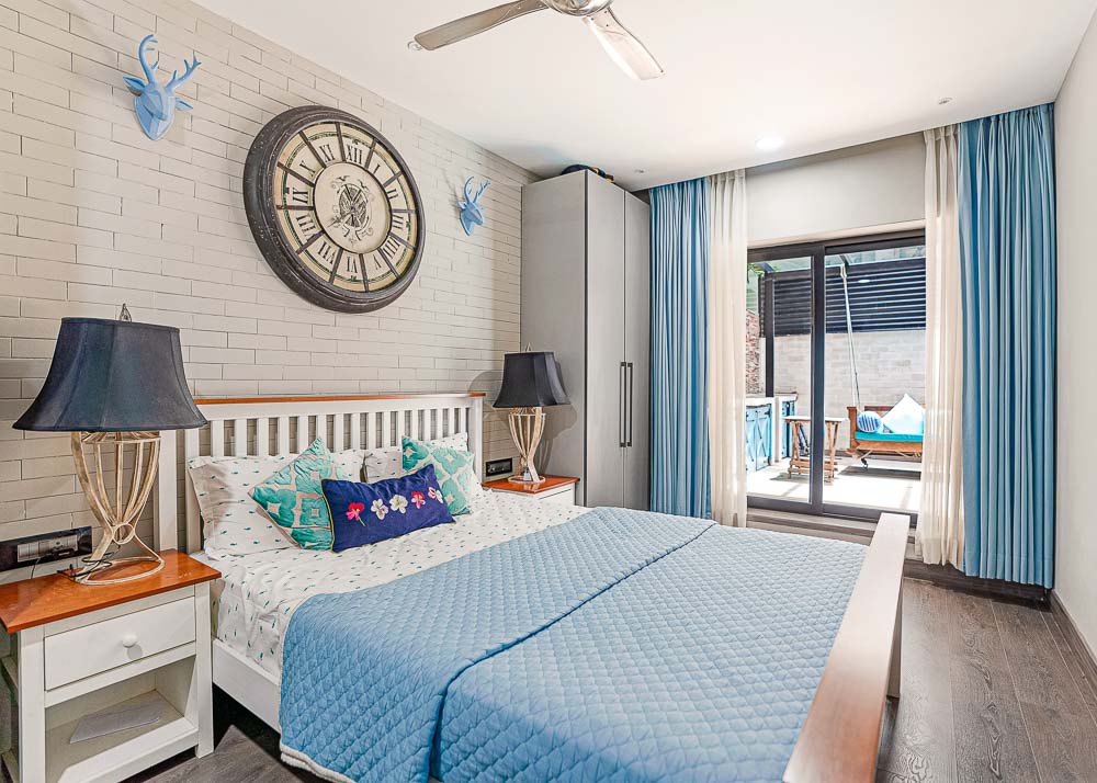 A bedroom with a white coloured wooden bed with a large clock on the wall, a couple of bedside tables, a wardrobe and a patio attached to it