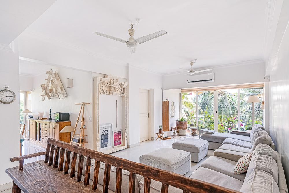 A living room with a beige sofa with the balcony at the end of it, overlooking a group of coconut trees