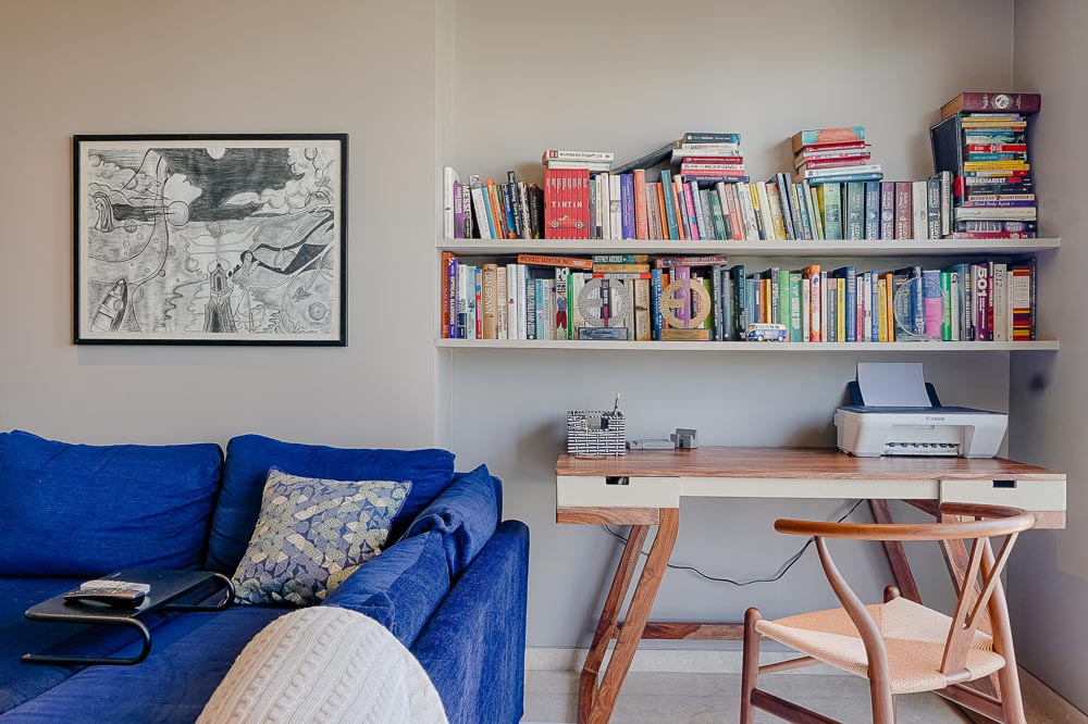 A colourful bookshelf hanging over a writing desk in wood, blue sofa and framed art on the wall