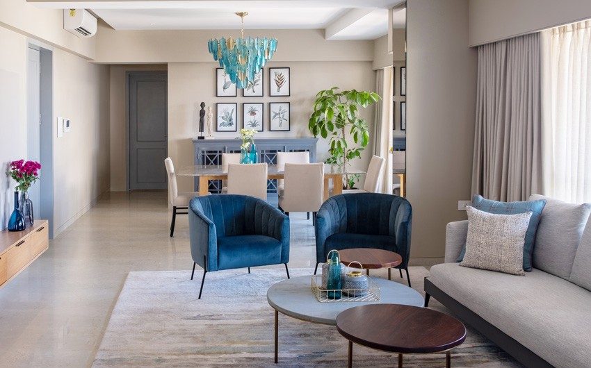 A living room with two chairs in blue fabric, an L shaped sofa and a dining table beyond the living room with a chandelier