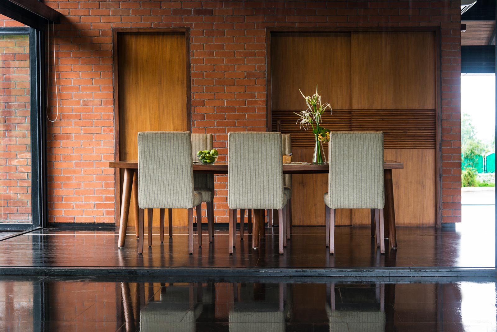Wooden dining table in forefront of brick wall, walls of windows on two sides, dark floor
