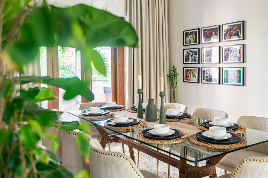 Dining table in glass and wood set with complete tableware, family photographs on the wall, bright sunlight from big window - Beautiful Homes