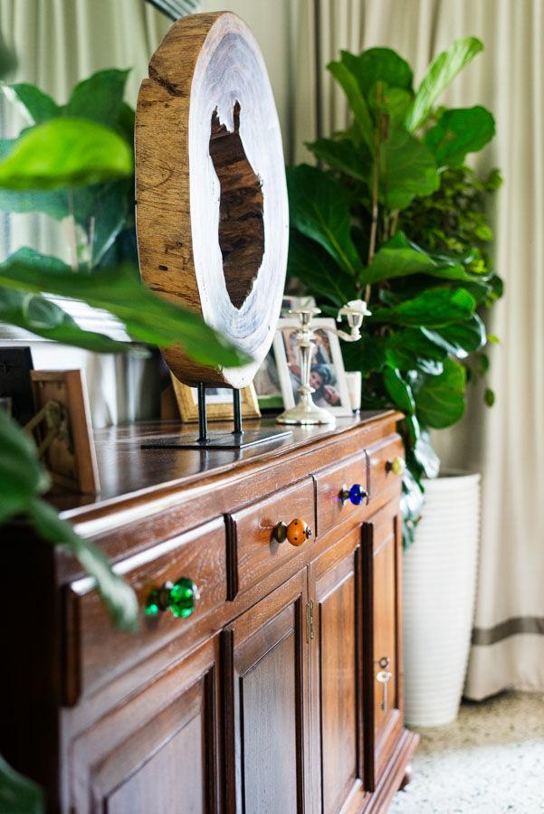 A wooden console with colourful knobs, frames kept on it and large potted plants on either side - Beautiful Homes