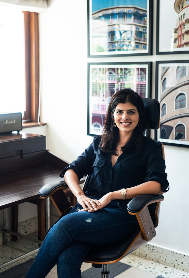Sarah Sham sitting on office armchair in leather, next to a wooden writing desk, framed photographs in the background