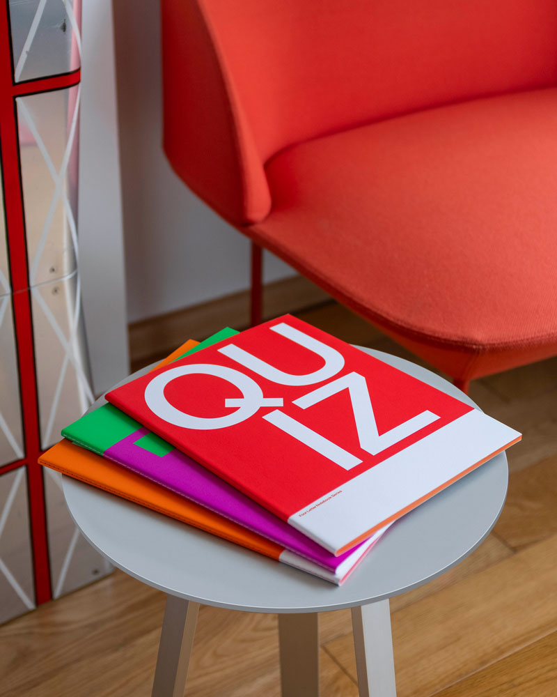 Three notebooks stacked together on a stool with 'Quiz' printed on the cover beside a red chair