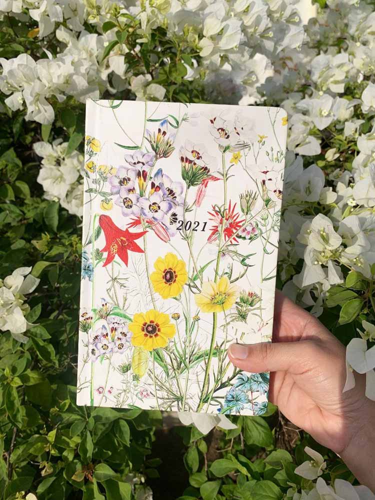 A diary with 2021 printed on it held by a hand in front of white bougainvilleas