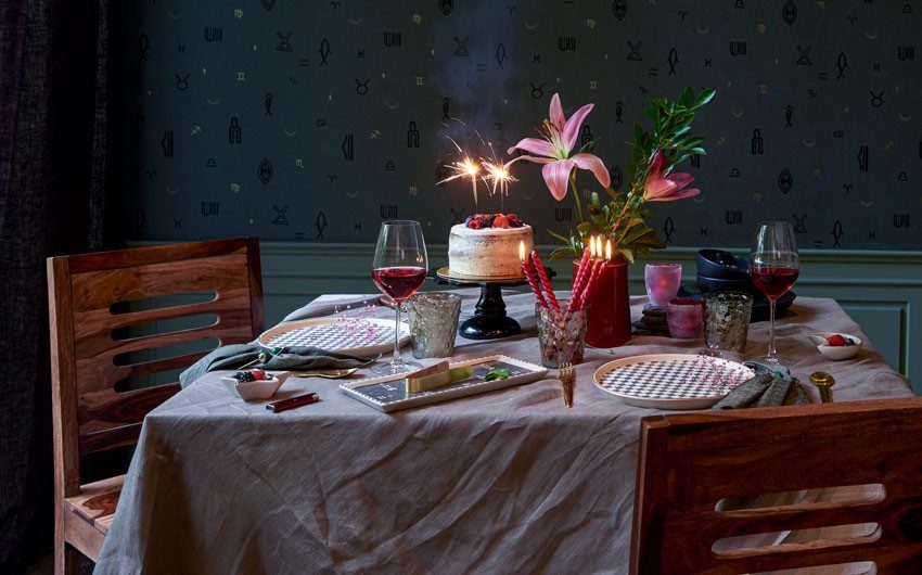 A 2 seater wooden dining table with a white table cloth, a cake, two plates, a wine glasses, a flower vase and lit candles in the centre