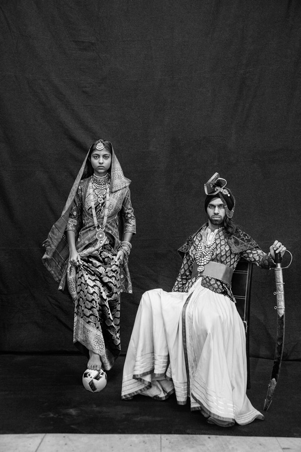 A man and a woman posing wearing Indian royal clothes, man sitting on a chair holding a sword and the woman with a football below her feet