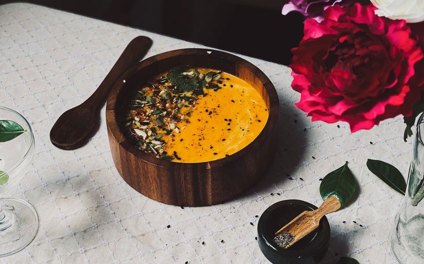 A wooden bowl filled with carrot soup placed on a table with a white coloured table cloth