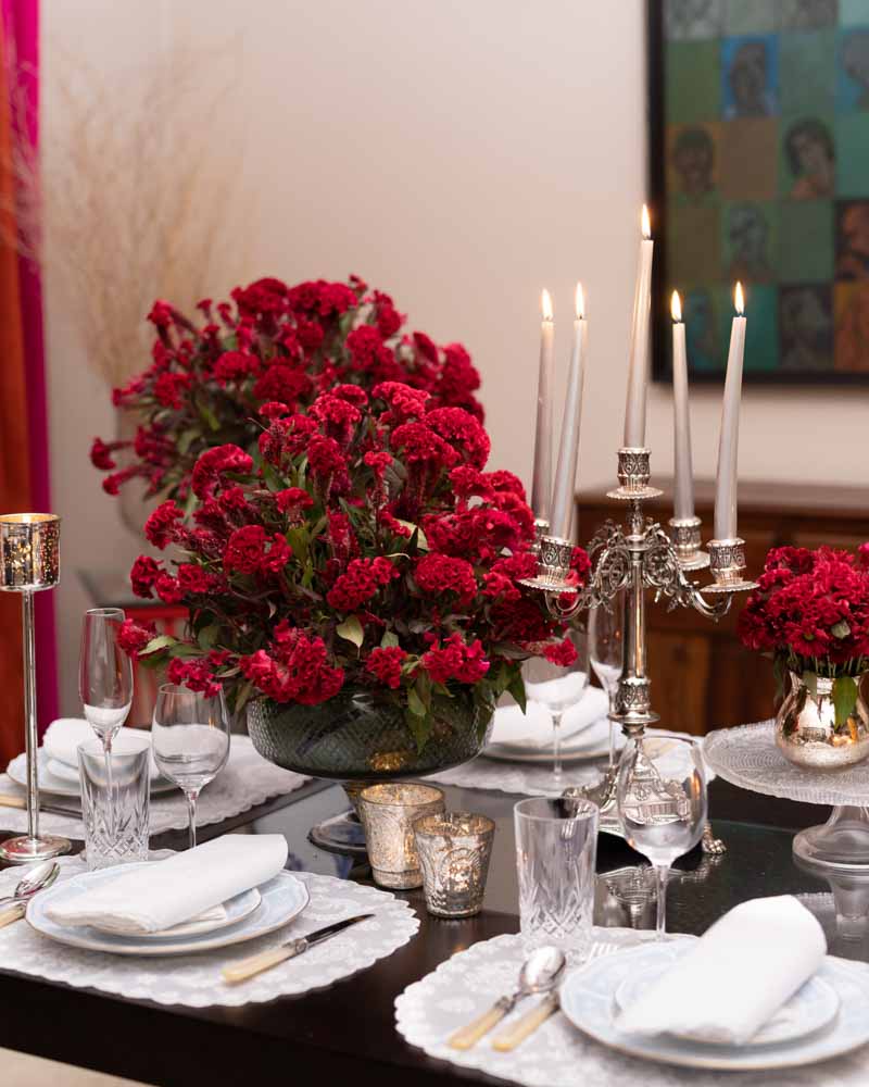 A table setting with red flowers, silver glasses and candle stands, and white ceramic plates