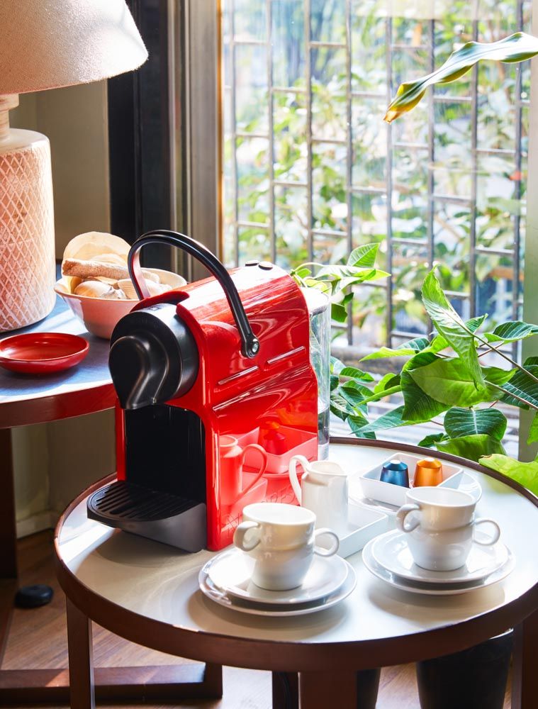 A small red coffee machine placed on a table beside a window along with espresso cups and coffee pods - Beautiful Homes