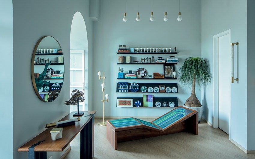 A room with five rows of shelves filled with different things like plates etc, a wooden lounger, a round mirror, a floor lamp and a planter